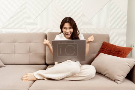 Téléchargez les photos : Excited Young Woman Celebrating Success On Laptop At Home, Sitting On Sofa, Expressing Joy And Happiness, Modern Interior - en image libre de droit