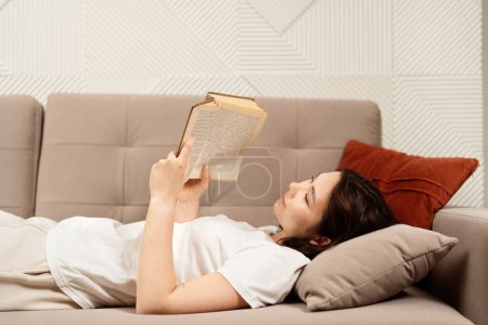 Relajación y ocio: Mujer joven disfrutando de un libro mientras descansa en un cómodo sofá, exhibiendo calma y satisfacción en casa