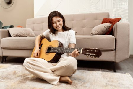 Fröhliche junge Frau spielt Gitarre drinnen, sitzt auf dem Boden neben dem Sofa im Wohnzimmer, entspannt Musik, genießt Freizeitaktivitäten zu Hause.