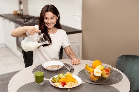 Mujer feliz vertiendo leche en el cereal en la cocina brillante, desayuno saludable, sonriendo, estilo de vida, interior moderno. Concepto de rutina diaria alegre y hábitos alimenticios saludables.