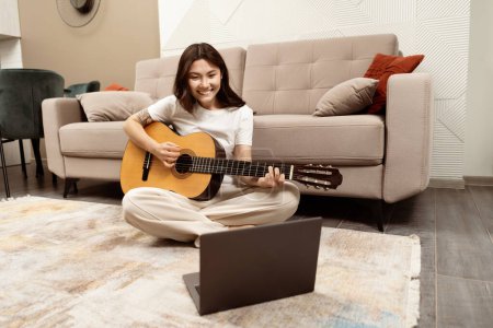 Photo for Joyful Young Woman Playing Guitar At Home, Sitting On Floor With Laptop Nearby, Expressing Creativity And Relaxation In Comfortable Modern Living Room. - Royalty Free Image