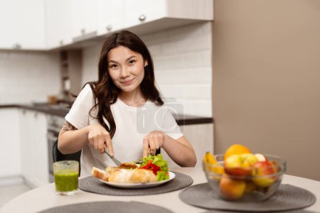 Téléchargez les photos : Femme souriante Manger des repas sains dans la cuisine moderne, mettant en vedette la salade fraîche et Croissant, mettant l'accent sur la nutrition et le mode de vie heureux - en image libre de droit