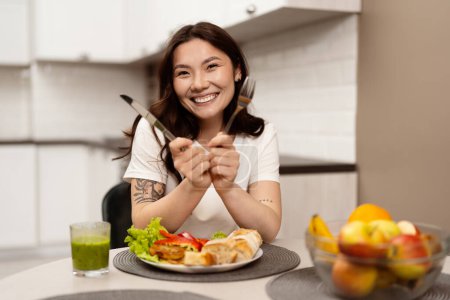 Joyeux jeune femme bénéficiant d'un repas sain dans une cuisine moderne, souriant joyeusement avec fourchette et couteau, aliments frais sur la table.