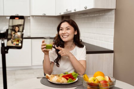 Fröhliche Frau nimmt gesundes Essen zu Hause auf, lächelnde weibliche Influencerin hält Smoothie in der Hand, demonstriert gesunden Lebensstil vor der Kamera, moderne Küche