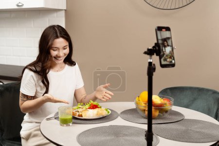 Food Blogger Creating Content: A cheerful young woman records a video of her healthy meal, showcasing cooking, lifestyle blogging, and digital content creation in a modern kitchen.