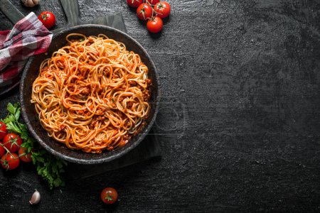 Spaghetti with Bolognese sauce in pan with napkin, tomatoes and garlic. On black rustic background Poster 657780544