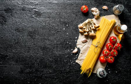 Photo pour Spaghettis crus sur papier avec champignons, épices et tomates sur une branche. Sur fond rustique noir - image libre de droit