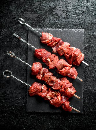 Foto de Pinzas con kebab crudo en una tabla de piedra. Sobre fondo rústico negro - Imagen libre de derechos