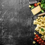 An assortment of different types of raw pasta with cheese, tomatoes and herbs. On dark rustic background