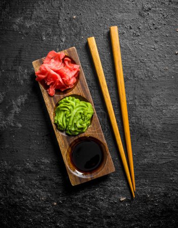 Foto de Wasabi, jengibre marinado y salsa de soja en un soporte de madera con palillos. Sobre fondo rústico negro - Imagen libre de derechos