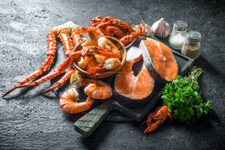 Fresh fish steak on a chopping Board with boiled shrimp, crayfish and crab. On black rustic background