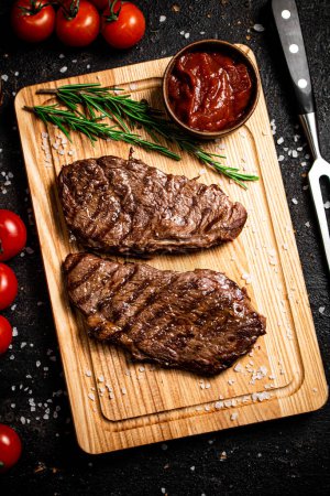 Téléchargez les photos : Steak grillé sur une planche à découper avec romarin et tomates. Sur fond noir. Photo de haute qualité - en image libre de droit