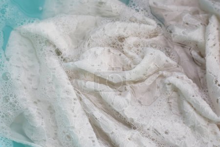 Téléchargez les photos : Robe en dentelle pour femme imbibée d'eau détergente dissoute avec bulle de mousse blanche. Concept de blanchisserie - en image libre de droit