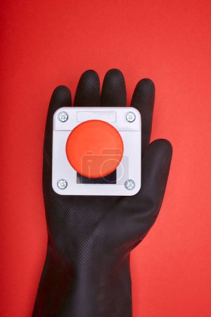 Photo for The hand in black resin glove, which keep the emergency button at the red background at the top view - Royalty Free Image