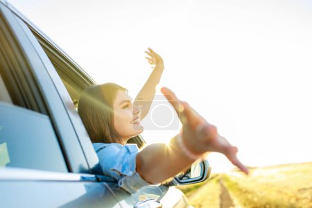 Mujer joven feliz mirando por la ventana del coche al atardecer dorado en el campo.
