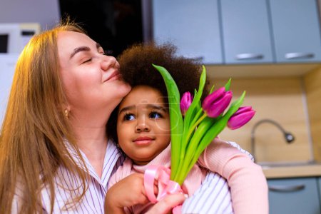 Foto de Madre poliétnica y su hija con peinado afro sentado en la cocina con un ramo de tulipanes y posrcard . - Imagen libre de derechos