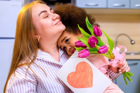Foto de Madre poliétnica y su hija con peinado afro sentado en la cocina con un ramo de tulipanes y posrcard . - Imagen libre de derechos