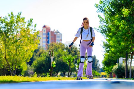 Porträt der schönen blonden Teenagerin mit coolen Zöpfen, weißem T-Shirt und ausgestellter pinkfarbener Hose beim Rollschuhlaufen im Park .