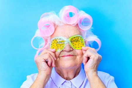 niedliche 80-jährige Frau mit Lockenwicklern auf weißem Haar trägt Hyaluronsäure-Augenmaske mit blauem Studiohintergrund.