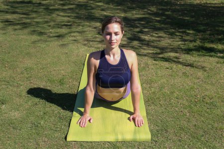 Téléchargez les photos : Jeune femme sportive pratiquant le yoga en plein air dans le parc par une belle journée ensoleillée. Yogini fait une variation de pose en updog. Fond de pelouse verte, espace de copie. - en image libre de droit