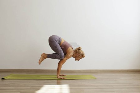 Sporty adult woman practicing crane yoga pose at home. Fit middle aged yogini doing the bakasana variation. White wall background, copy space, close up.