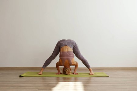 Sporty adult woman practicing hatha yoga at home. Fit middle aged yogini doing the wide legged forward bend pose. White wall background, copy space, close up.
