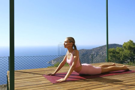 Photo for Young woman practicing hatha yoga on a terrace with a beautiful ocean an mountains view. Yogini doing the upward facing dog pose. Copy space, background. - Royalty Free Image