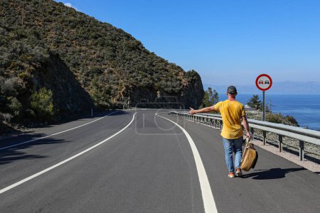 Photo for Traveling musician with a guitar walking alone down the winding road with a beautiful sea view, trying to stop a car by showing a hitchhiking gesture. - Royalty Free Image