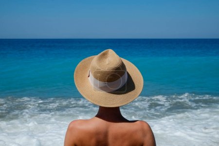 Foto de Vista posterior de una mujer joven con sombrero de paja de ala ancha, disfrutando de la vista al mar. Fondo, primer plano, espacio de copia - Imagen libre de derechos