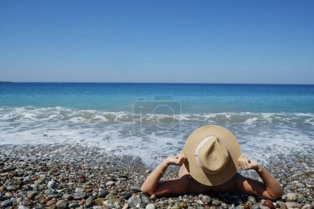 Foto de Vista posterior de una joven que cubre su cuerpo con sombrero de paja de ala ancha. Mujer tomando el sol en la orilla del océano atlántico. Cerrar, copiar espacio, fondo. - Imagen libre de derechos