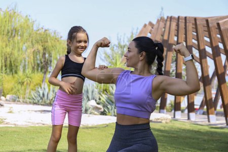 Téléchargez les photos : Super maman fléchissant son puissant biceps avec sa fille. Mère sportive et sa petite fille se lient par l'exercice physique. Gros plan, espace de copie, fond. - en image libre de droit