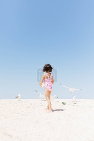 Foto de Niña feliz vistiendo traje de baño alimentando gaviotas en el día de playa. Fotografía de estilo de vida. Colores pastel - Imagen libre de derechos