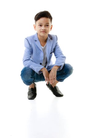 Téléchargez les photos : Un garçon asiatique de 10 ans vêtu d'une veste décontractée est assis intelligent et heureux en regardant la caméra sur un fond blanc isolé. Concept de style de vie des enfants et publicité générale. - en image libre de droit