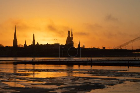 Téléchargez les photos : Un beau paysage de lever de soleil coloré avec silhouette de bâtiment du centre-ville contre un ciel matinal lumineux. Un paysage hivernal avec rivière gelée Daugava dans la capitale de la Lettonie Riga. - en image libre de droit