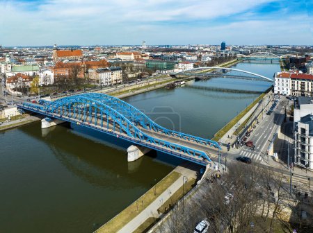Bridges on Vistula River in Krakow, Poland. Aerial view. Boulevards with waking people. Blue tied-arc bridge in front. Footbridge for pedestrians and cyclist. Far view of new double railway bridge