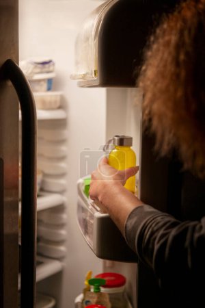 Photo for Woman hand by the open refrigerator at night outstretched to a bottle of drink - Royalty Free Image