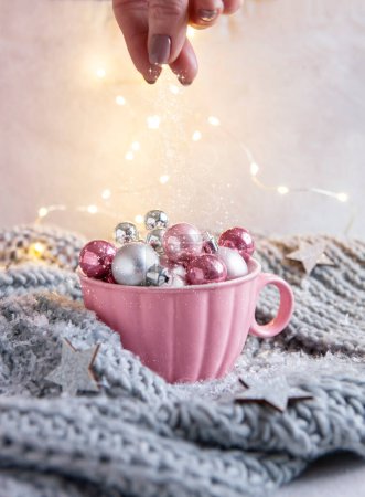 Téléchargez les photos : Composition magique de Noël et du Nouvel An, boules de Noël dans une grande tasse rose et main d'une femme jetant de la neige. Mise au point sélective, gros plan, fond - en image libre de droit