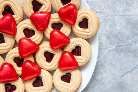 Foto de Primer plano de galletas en forma de corazón y chocolates en envoltura roja en el plato. Fondo, vista superior, espacio para copiar - Imagen libre de derechos