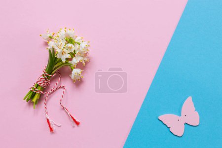 Photo for Fresh beautiful bouquet of the first spring forest snowdrops with a red and white martisor cord and a butterfly figure - a traditional symbol of the first spring day on a pink pastel and blue paper background - Royalty Free Image