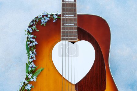 Photo for Acoustic guitar with beautiful blue spring forgot-me-not flowers and white heart shape silhouette on blue sky background. - Royalty Free Image