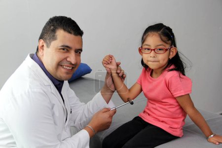 Foto de Latino doctor medic and girl patient in medical office checks her reflexes on hammer in her checkup to find disease diagnosis - Imagen libre de derechos