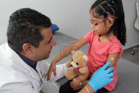 Dark-haired Latino doctor and little girl have a medical consultation in the pediatric office to vaccinate their arm against Covid, chickenpox, diphtheria, influenza, hepatitis, measles, mumps