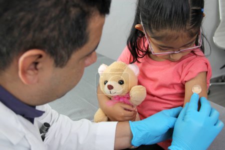 Dark-haired Latino doctor and little girl have a medical consultation in the pediatric office to vaccinate their arm against Covid, chickenpox, diphtheria, influenza, hepatitis, measles, mumps