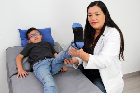 Foto de Médico pediatra latina atiende a un niño de 7 años en su consultorio revisa su pierna, rodilla y pie en busca de caídas y enfermedades como raquitismo, blount, tedinitis, rotura o lesión - Imagen libre de derechos