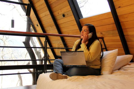 Téléchargez les photos : Femme latine de 40 ans travaille avec son ordinateur portable sur le lit d'une cabane au milieu de la forêt en tant que nomade numérique - en image libre de droit