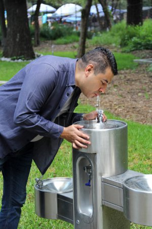 Dark-haired 40-year-old Latino man drinks drinking water from a public drinking fountain in an open space on the street due to the extreme heat wave