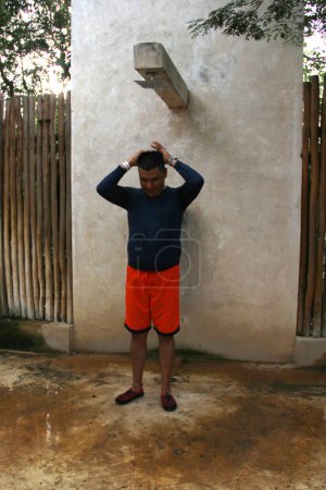 Dunkelhäutiger erwachsener Mann mit Latino-Badeanzug und Aqua-Schuhen badet und spült in öffentlichen Wellnessduschen, um Sand und Sonnencreme zu entfernen
