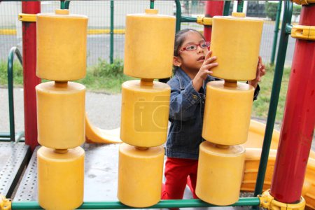 Téléchargez les photos : 4 ans Latina fille brune avec des lunettes joue dans le parc jeux dans la solitude et la pauvreté passe ses jours de vacances avec l'autisme - en image libre de droit