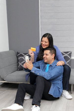 Couple of Latin man and woman sitting in their living room using cell phone and credit card to pay, buy with discounts and promotions