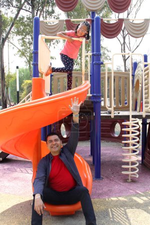 Divorced single dad plays with his brunette Latina daughter on the park slide enjoying quality time together celebrating Father's Day